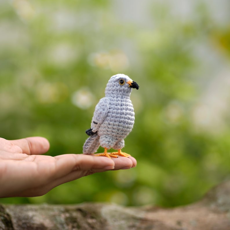 May include: A small, handcrafted crochet bird figurine featuring light gray yarn, a black beak, dark eyes, and bright orange feet. The bird is designed with intricate stitching to mimic feathers and stands upright on a flat surface.