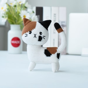 May include: A crocheted calico cat toy with brown, black, and white fur. The cat is standing on a white surface.