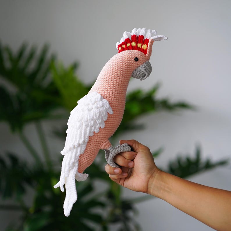 以下が含まれることがあります： ピンク、白、赤、グレーの色合いの手作りの鉤針編みのオウム。鳥は特徴的なとさかと白い翼を持っています。オウムは手で持たれています。かぎ針編みの鳥は装飾品です。