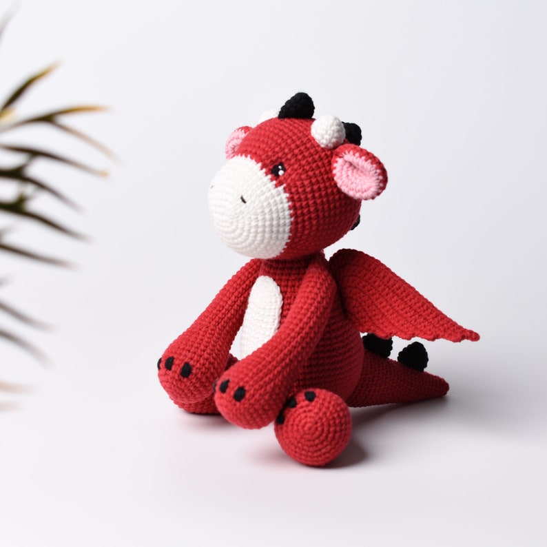 May include: A red crocheted dragon toy with black eyes and a white belly. The dragon is sitting with its wings spread out and has a small red crocheted ball in front of it.