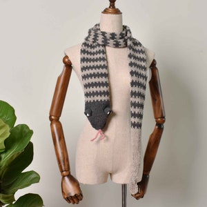 May include: A gray and beige striped knitted scarf with a snake head design. The snake has black eyes and a pink tongue.