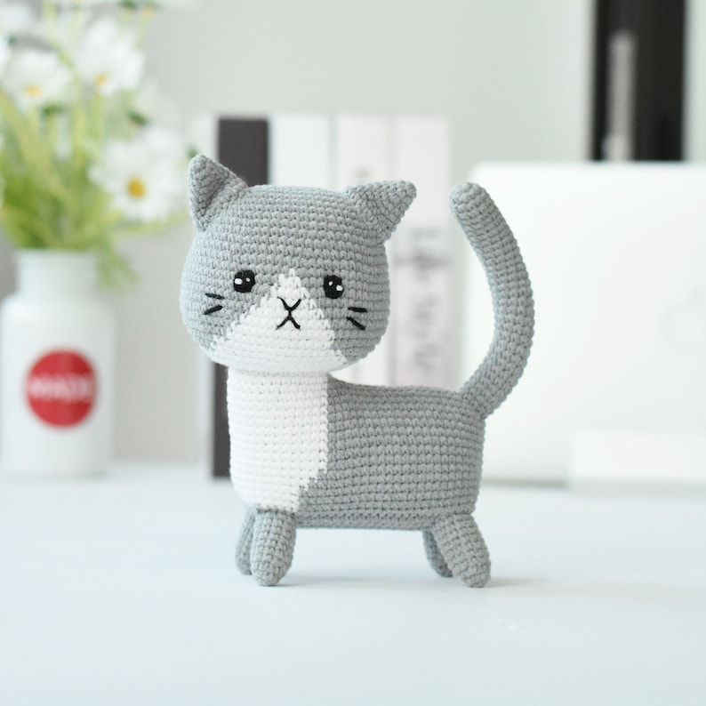 May include: A gray and white crocheted cat toy with a white face and black eyes. The cat is standing on a white surface.