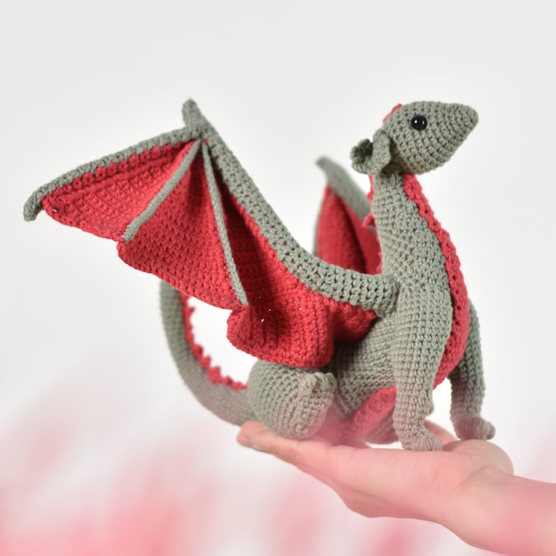 May include: A gray crocheted dragon with red wings. The dragon is sitting on a hand.