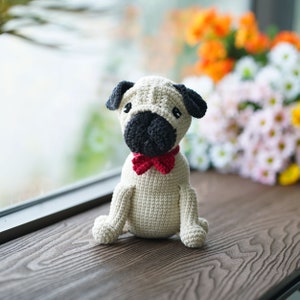 Puede incluir: Un juguete de perro pug de crochet con una pajarita roja. El juguete está sentado sobre una superficie de madera.
