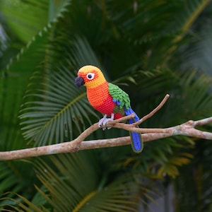 May include: A crocheted parrot toy with red, orange, yellow, green, and blue colors. The parrot is perched on a branch with green leaves in the background.