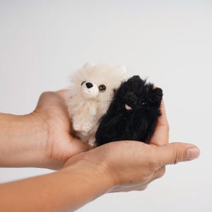 Puede incluir: Dos pequeños peluches de perro Pomerania. Uno es de color crema, el otro es negro. Los juguetes tienen ojos y narices pequeños y oscuros, y el negro tiene una lengua rosada. Los juguetes se sostienen en manos ahuecadas.