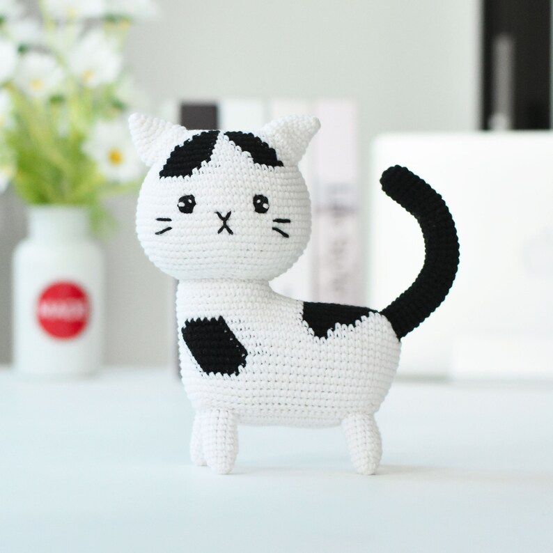 May include: A white crocheted cat with black spots and a black tail. The cat has a cute face with black eyes and a black nose.