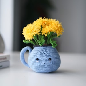 May include: A handmade blue crochet vase with a smiling face and a handle, filled with yellow crochet dandelions and green stems. The vase is sitting on a white surface.