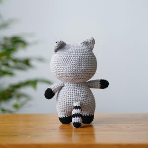 May include: A grey crocheted raccoon plush toy with black stripes on its tail and black accents on its ears and paws.