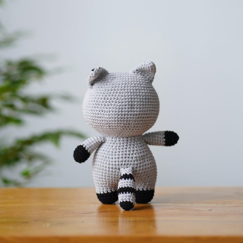 May include: A grey crocheted raccoon plush toy with black stripes on its tail and black accents on its ears and paws.