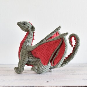May include: A crocheted dragon toy with green and red yarn. The dragon has a long tail, wings, and a small head with black eyes.