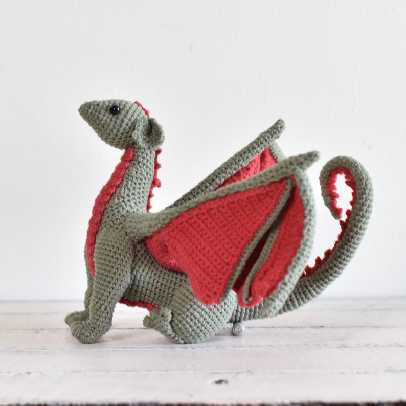May include: A crocheted dragon toy with green and red yarn. The dragon has a long tail, wings, and a small head with black eyes.