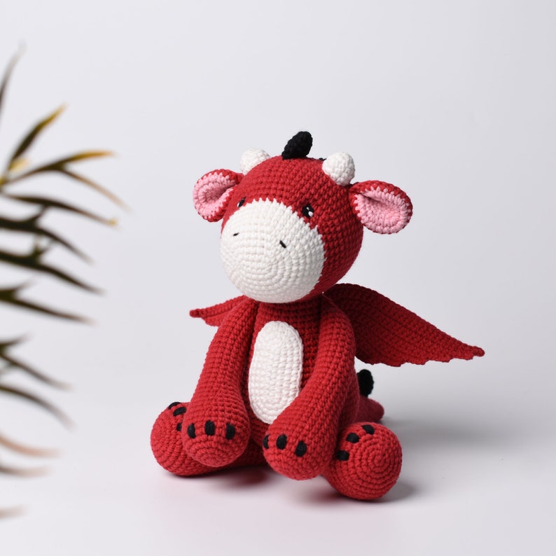 May include: A red crocheted dragon toy with white accents and black eyes. The dragon is sitting with its wings spread out.