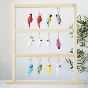 Puede incluir: Un expositor de madera exhibe una colección de llaveros de loros de ganchillo. Los llaveros presentan varios colores, incluyendo rojo, azul, amarillo y blanco. Cada loro tiene un llavero plateado.