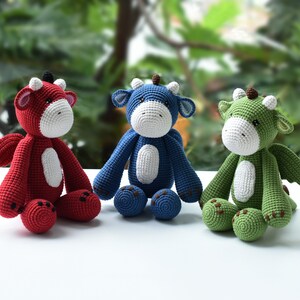 May include: Three crocheted dragon plushies, one red, one blue, and one green, sitting on a white surface. Each plush has a white belly and brown eyes.
