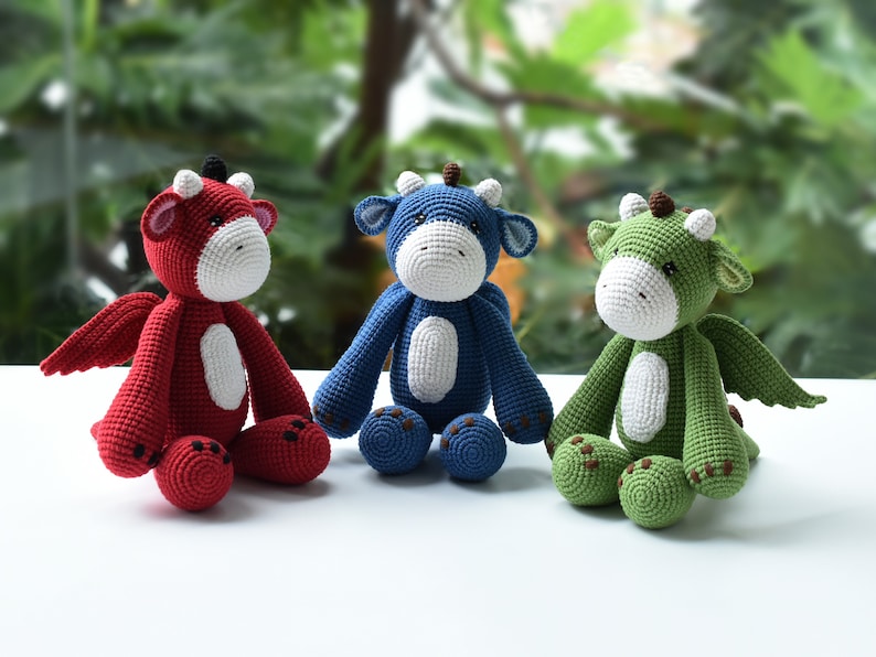 May include: Three crocheted dragon plushies, one red, one blue, and one green, sitting on a white surface. Each plush has a white belly and brown eyes.