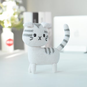 May include: A white crocheted cat with gray stripes. The cat has a black embroidered nose and eyes.