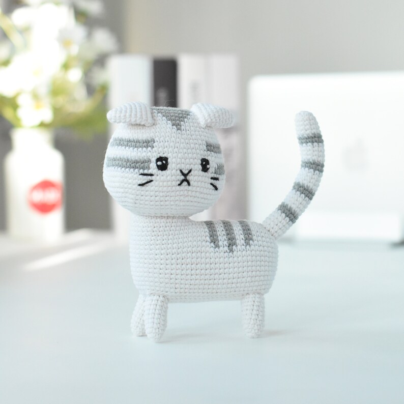 May include: A white crocheted cat with gray stripes. The cat has a black embroidered nose and eyes.