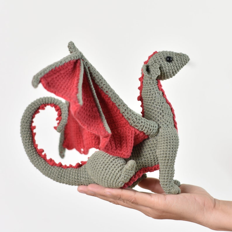 May include: A gray crocheted dragon with red wings. The dragon is sitting on a hand.