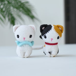 May include: Two crocheted cat toys. One is white with a blue scarf and the other is white with black and orange patches and a red collar.