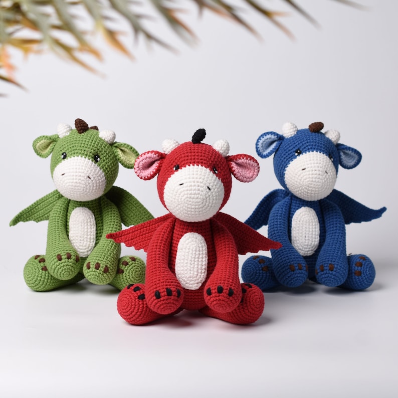 May include: Three crocheted dragon toys, one green, one red, and one blue, sitting on a white surface. Each dragon has a white face and black eyes.