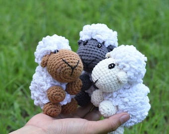 May include: Three crocheted sheep plushies in different colors: white, brown, and gray. The sheep are sitting on a hand.