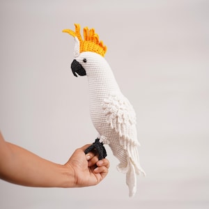 以下が含まれることがあります： 黄色い冠と黒い嘴と足を持つ、手作りの白いかぎ針編みのオウム。鳥は手に止まっており、アミグルミデザインの詳細な職人技を紹介しています。鳥の羽は複雑にかぎ針編みされています。