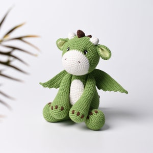 May include: A green crocheted dragon toy with white accents and brown details. The dragon is sitting with its wings out and has a cute expression.