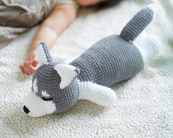 May include: A gray and white crocheted husky dog plush toy lying on a white fuzzy blanket.