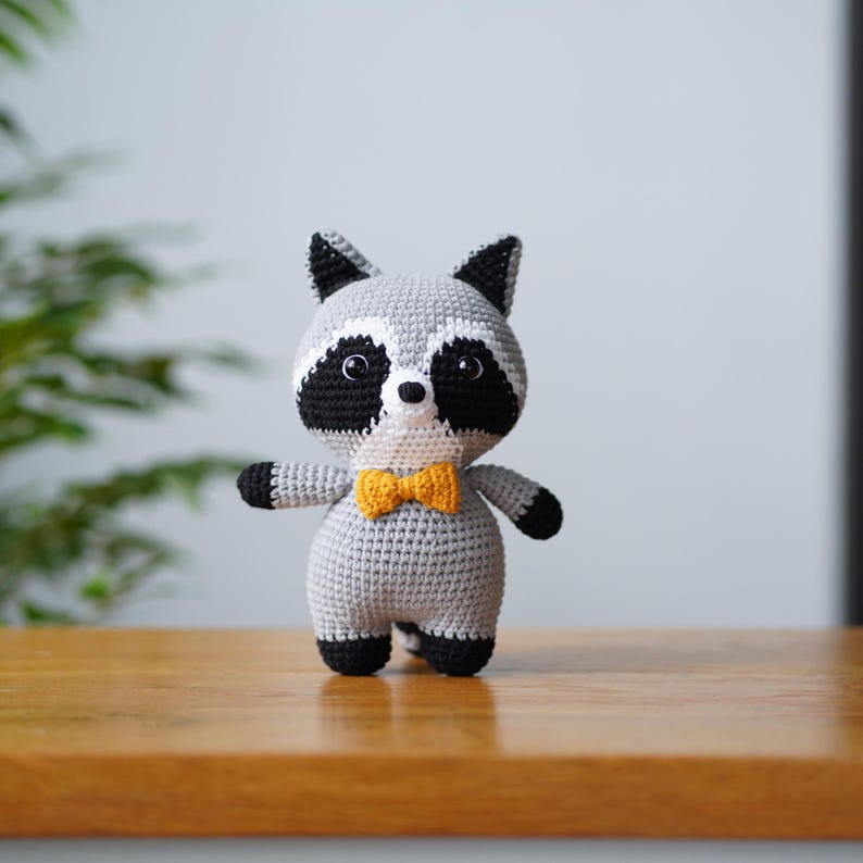 May include: A grey crocheted raccoon plush toy wearing a yellow bow tie. The toy has black eyes and a white nose.