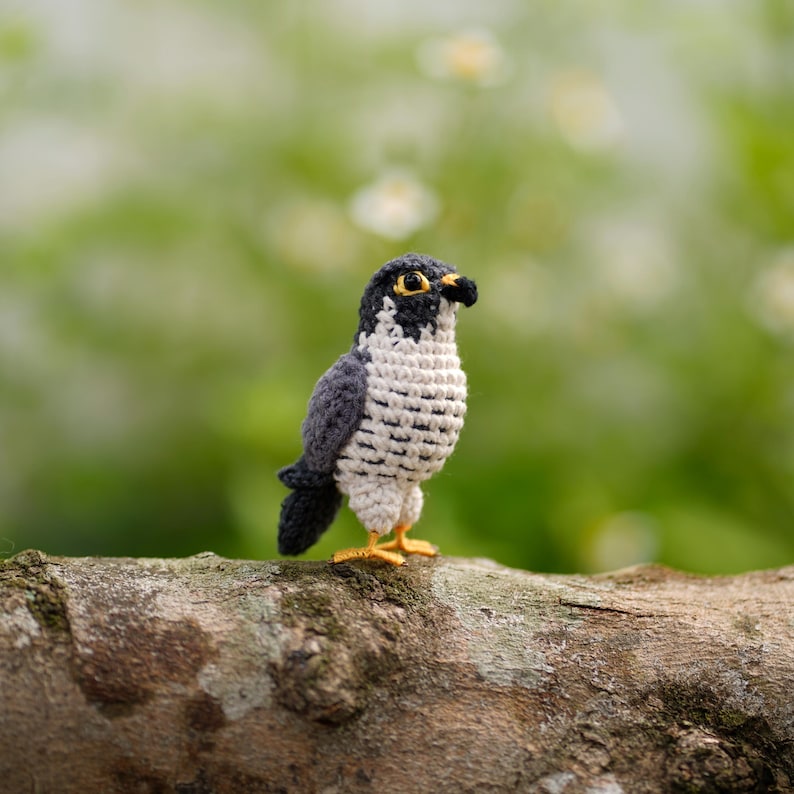 May include: A small, handcrafted crochet falcon figurine featuring dark gray wings, a white chest with black horizontal stitching, a black head with a yellow eye, and a small black beak. The bird stands on two bright yellow yarn feet.