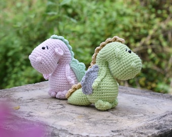 Puede incluir: Dos juguetes de peluche de dragón hechos a crochet. Uno es rosa y el otro es verde. Están sentados sobre una superficie de hormigón gris.