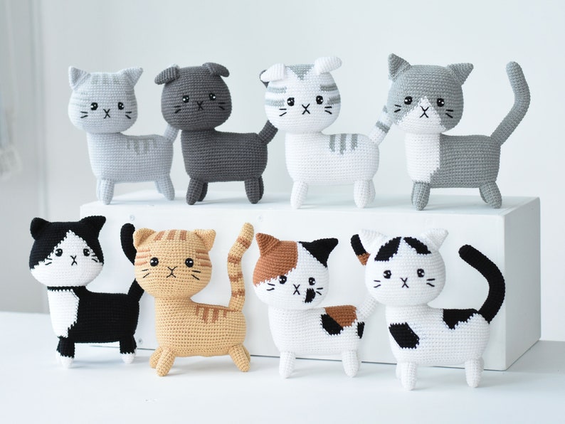 May include: Eight crocheted cat plushies in various colors and patterns, including black and white, gray and white, brown and white, and orange and white. The plushies are arranged in two rows on a white surface.