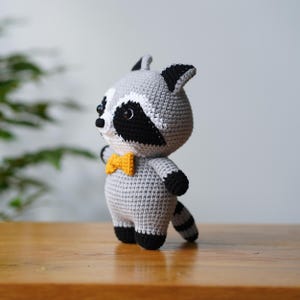 May include: A crocheted raccoon plush toy with a grey body, black eyes, and a black nose. The toy is wearing a yellow bow tie and has a black tail.