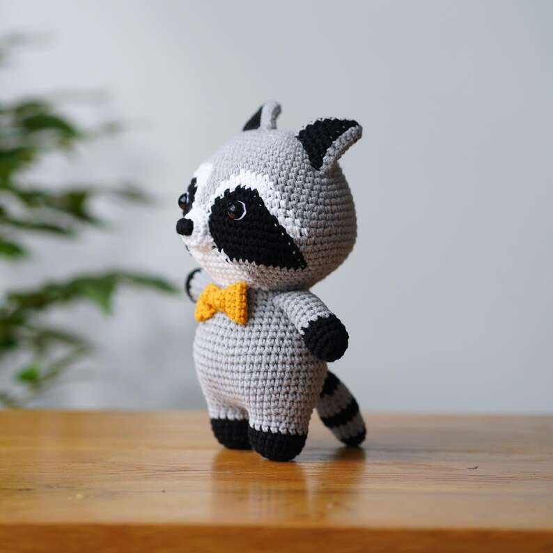 May include: A crocheted raccoon plush toy with a grey body, black eyes, and a black nose. The toy is wearing a yellow bow tie and has a black tail.