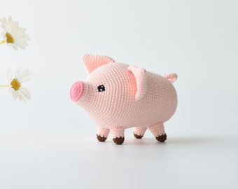 May include: A pink crocheted pig toy with black eyes and a pink snout. The pig is standing on a white surface.