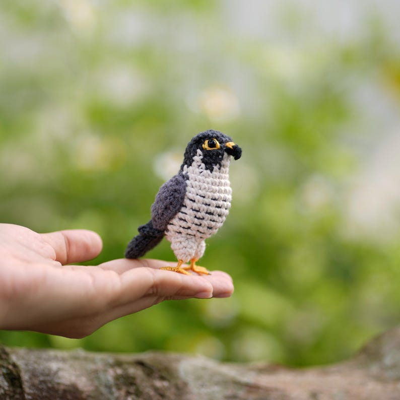 May include: A small, handcrafted crochet falcon figurine featuring a dark gray back and wings, a cream-colored chest with dark gray horizontal markings, a black head with a distinct facial pattern, yellow eyes, and small yellow feet.