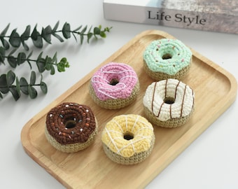 Puede incluir: Cinco donuts de crochet en una bandeja de madera. Los donuts son de varios colores, incluyendo rosa, verde, marrón, amarillo y blanco. Cada donut tiene una cobertura diferente, como chispas, chispas de chocolate o glaseado blanco.