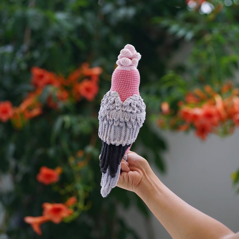 以下が含まれることがあります： ピンク、グレー、黒色の手編みのかぎ針編み鳥。 鳥はピンクの頭と胸、灰色の翼、黒い尾を持っています。 緑の葉とオレンジ色の花を背景に、手で持っています。