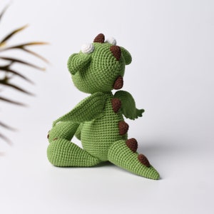 May include: Green crocheted dragon toy with brown spots and white eyes. The dragon is sitting on its hind legs with its wings spread out.
