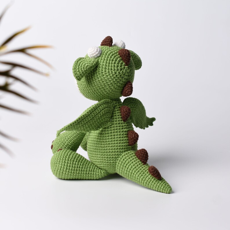 May include: Green crocheted dragon toy with brown spots and white eyes. The dragon is sitting on its hind legs with its wings spread out.