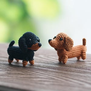 Könnte beinhalten: Zwei gehäkelte Dackel-Hundefiguren. Eine ist schwarz mit braunen Akzenten, die andere ist braun. Sie befinden sich auf einer Holzoberfläche mit einem verschwommenen grünen Hintergrund. Die Hunde sind handgefertigt und haben kleine, detaillierte Merkmale.