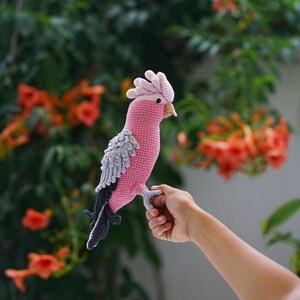 以下が含まれることがあります： ピンク、グレー、黒色の手編みの鳥。鳥はピンク色の体、灰色の羽、黒色の尾を持っています。手の上に止まっており、背景には緑の葉とオレンジ色の花があります。