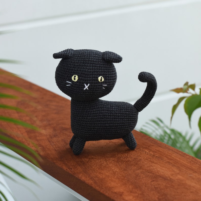 May include: A black crocheted cat toy with yellow eyes and white whiskers. The cat is standing on a wooden surface.