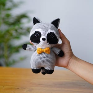 May include: A grey crocheted raccoon plush toy wearing a yellow bow tie. The toy has black eyes and a white nose.