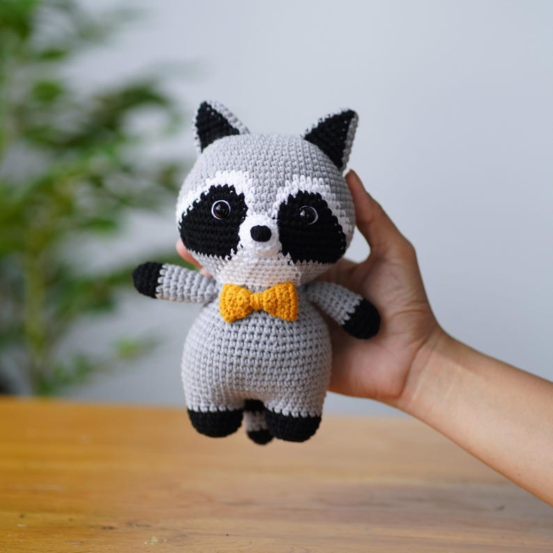 May include: A grey crocheted raccoon plush toy wearing a yellow bow tie. The toy has black eyes and a white nose.