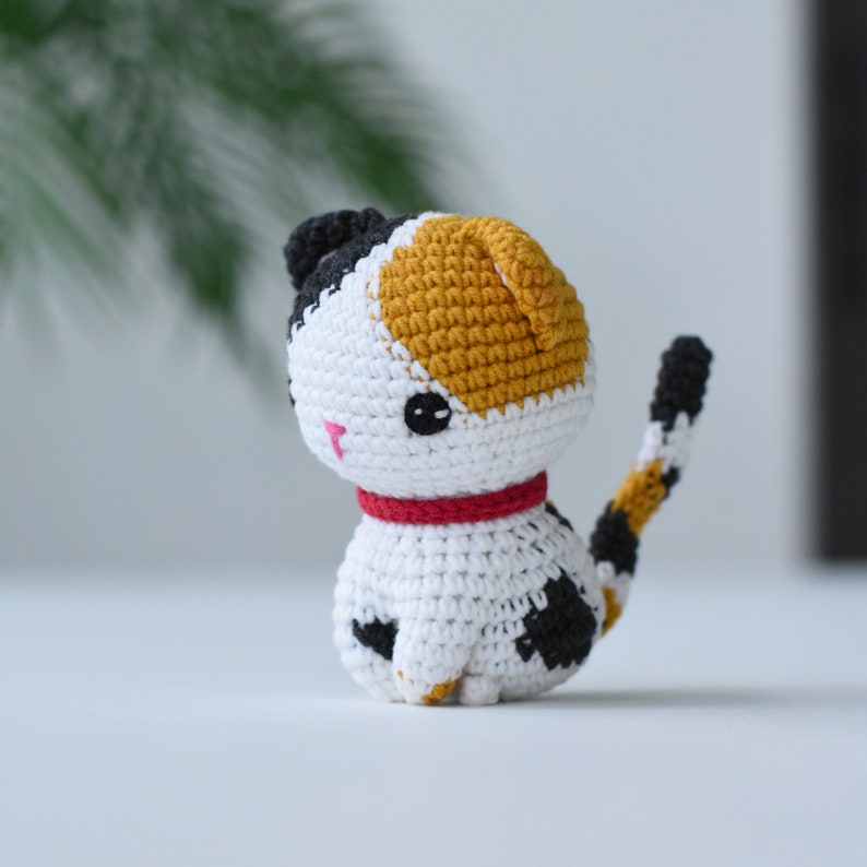 May include: A crocheted calico cat toy with black, white, and orange yarn. The cat has a red collar and black eyes.