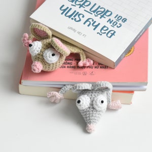 May include: Two crocheted mouse bookmarks, one brown and one gray, with large white eyes and pink noses. The bookmarks are placed on top of a stack of books.