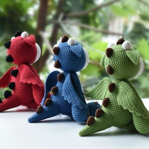 May include: Three crocheted dragon toys, one red, one blue, and one green, sitting on a white surface. The dragons are facing away from the camera, with their wings spread out.