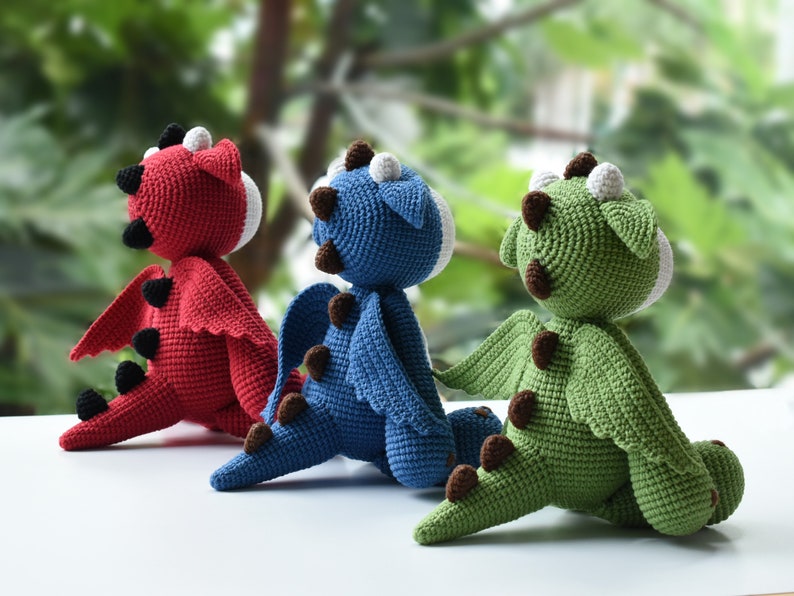 May include: Three crocheted dragon toys, one red, one blue, and one green, sitting on a white surface. The dragons are facing away from the camera, with their wings spread out.